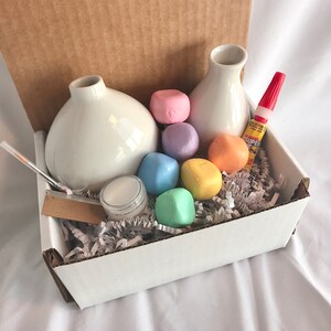 May include: A white box filled with craft supplies for making a vase. The box contains two white ceramic vases, a small jar of glue, a red glue stick, and a variety of pastel-colored foam balls.