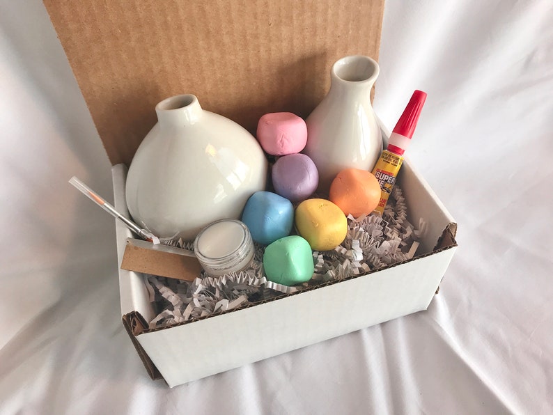 May include: A white box filled with craft supplies for making a vase. The box contains two white ceramic vases, a small jar of glue, a red glue stick, and a variety of pastel-colored foam balls.