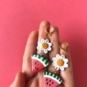 May include: A pair of dangle earrings featuring a watermelon slice and a white daisy charm. The watermelon slice is red, green, and black, and the daisy has a yellow center.