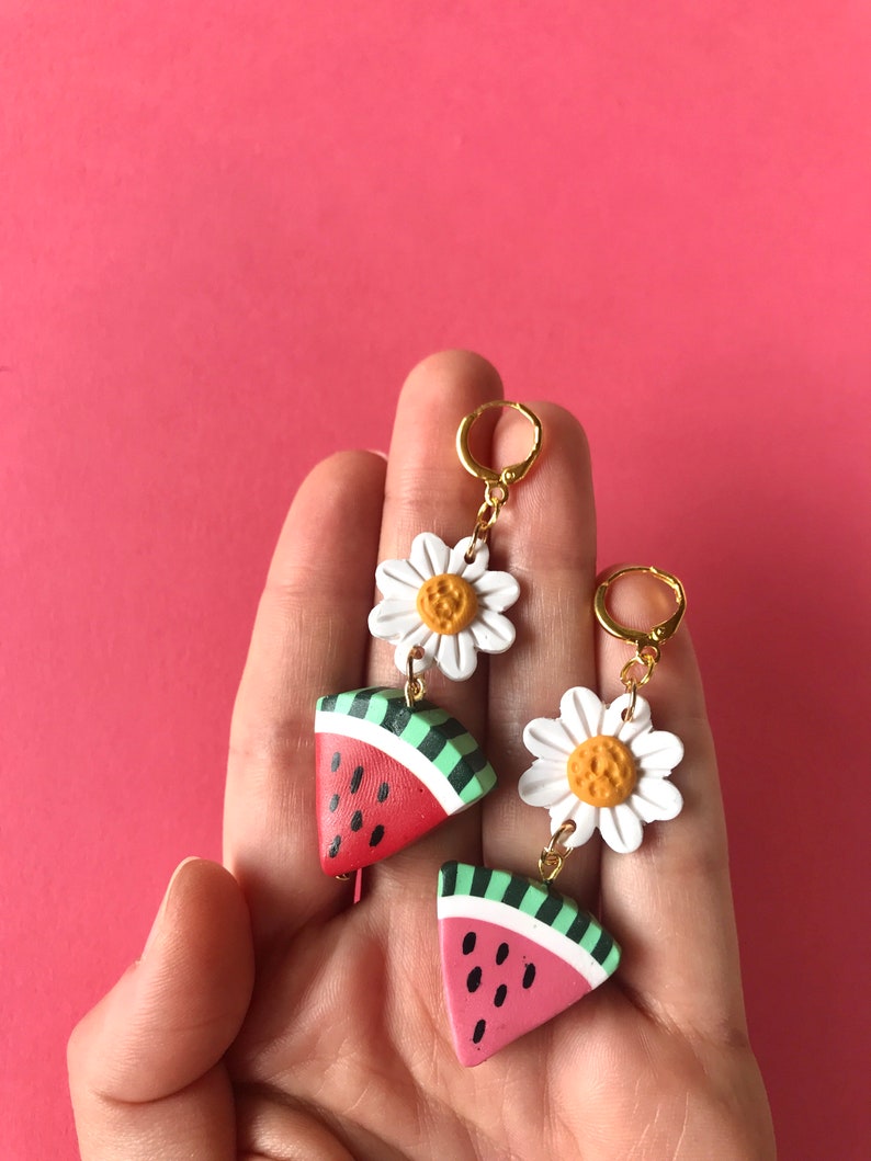May include: A pair of dangle earrings featuring a watermelon slice and a white daisy charm. The watermelon slice is red, green, and black, and the daisy has a yellow center.