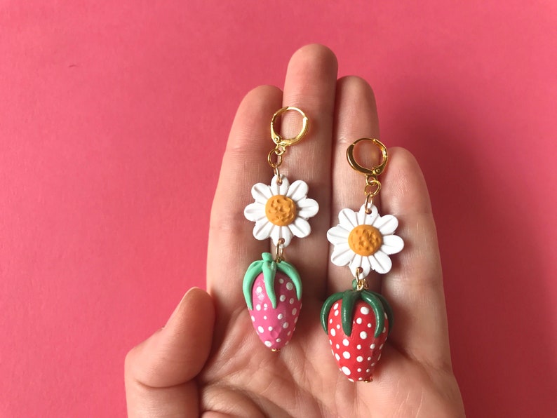 May include: A pair of dangle earrings featuring a white daisy with a yellow center and a red and white strawberry with green leaves.