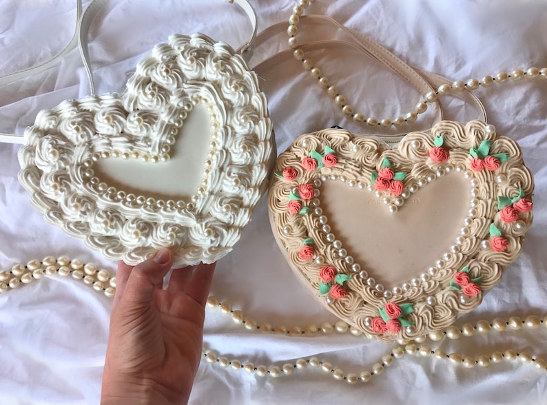 May include: Two heart-shaped purses with a cream-colored background and pearl accents. One purse has a white frosting-like design and the other has pink flower accents.