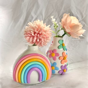 May include: Two ceramic vases with pastel rainbow and flower designs. The vase on the left has a rainbow design with pastel pink, yellow, orange, green, blue, and purple colors. The vase on the right has a white background with pastel pink, yellow, orange, blue, and purple flowers.