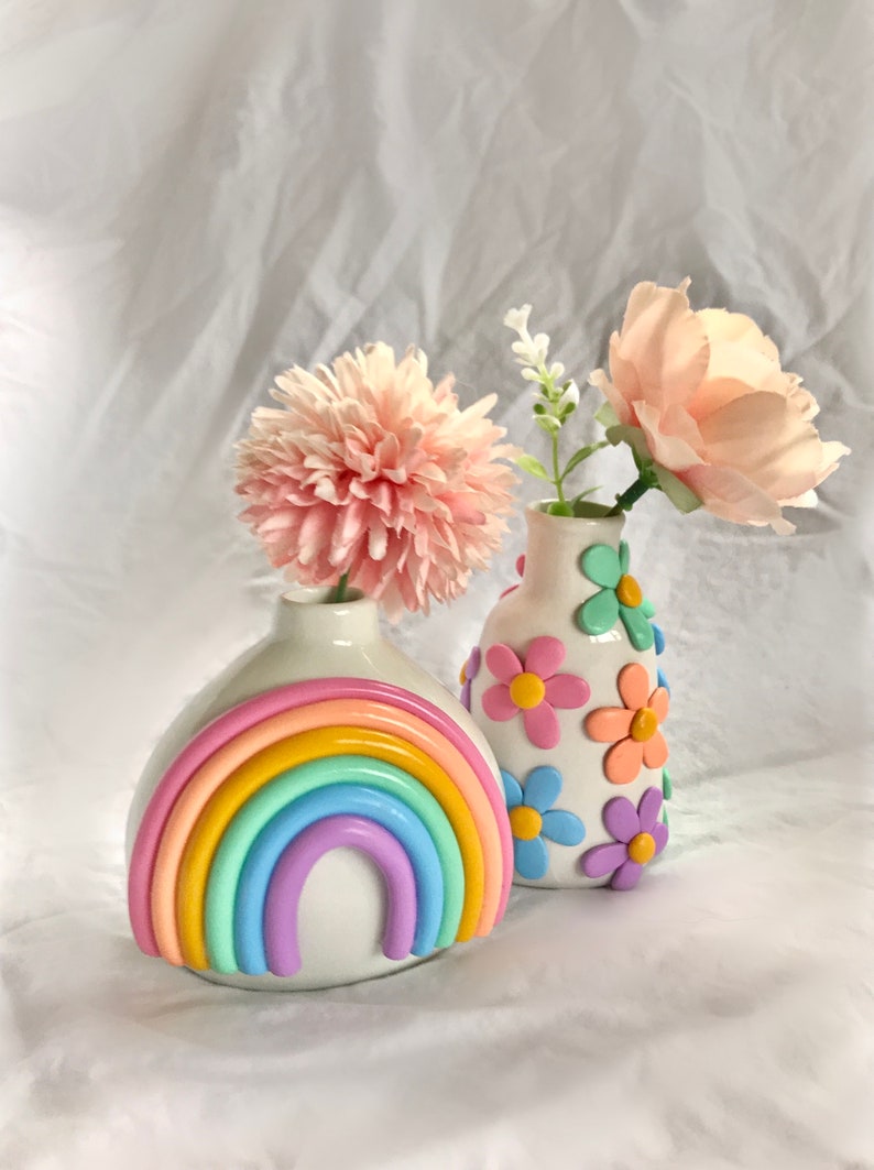 May include: Two ceramic vases with pastel rainbow and flower designs. The vase on the left has a rainbow design with pastel pink, yellow, orange, green, blue, and purple colors. The vase on the right has a white background with pastel pink, yellow, orange, blue, and purple flowers.