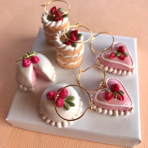 May include: Four pairs of dangle earrings featuring miniature cake and heart shapes with pink and white frosting and strawberry accents. The earrings are made of polymer clay and gold hoops.