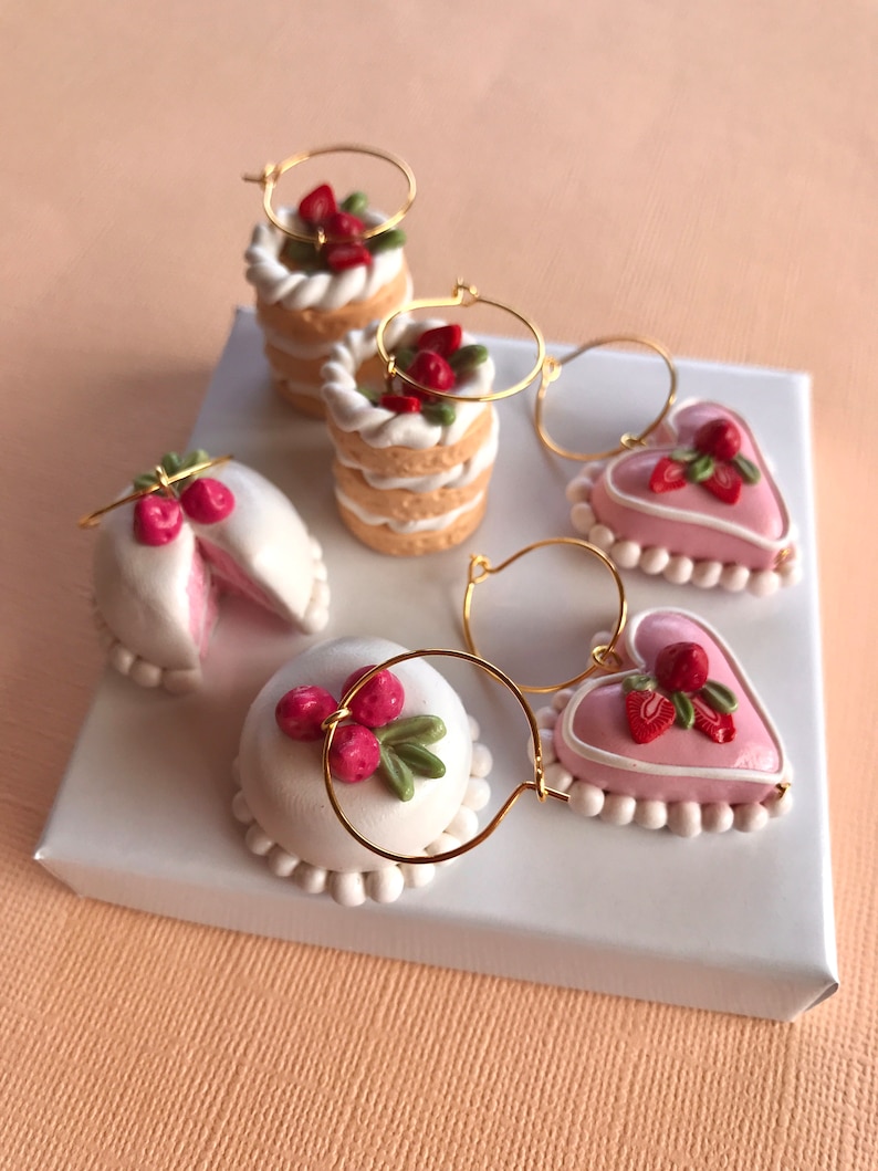 May include: Four pairs of dangle earrings featuring miniature cake and heart shapes with pink and white frosting and strawberry accents. The earrings are made of polymer clay and gold hoops.