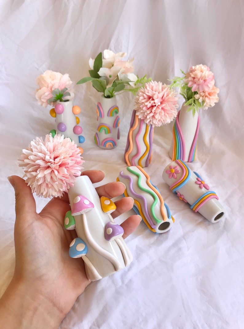 May include: A set of seven small ceramic vases with different pastel-colored designs. The vases are decorated with rainbows, wavy lines, dots, and mushrooms. The vases are perfect for displaying small bouquets of flowers.