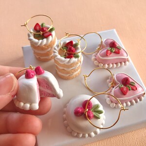 May include: Five pairs of handmade polymer clay earrings shaped like cakes and hearts with strawberry and green leaf accents. The earrings are on a white surface.