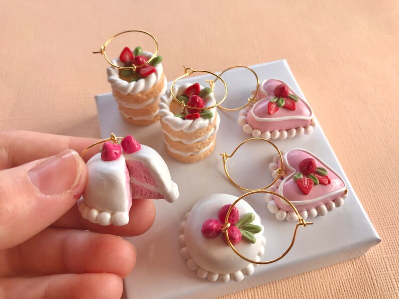 May include: Five pairs of handmade polymer clay earrings shaped like cakes and hearts with strawberry and green leaf accents. The earrings are on a white surface.