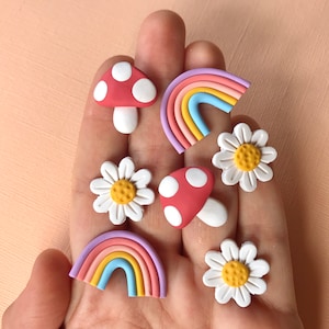 May include: A set of six colorful clay earrings featuring a rainbow, mushroom, and daisy designs. The earrings are handmade and have a whimsical, retro aesthetic.