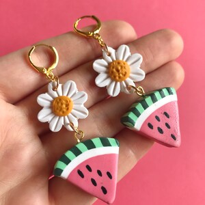 May include: A pair of dangle earrings featuring a white daisy charm and a pink watermelon slice charm. The charms are connected by a gold chain.