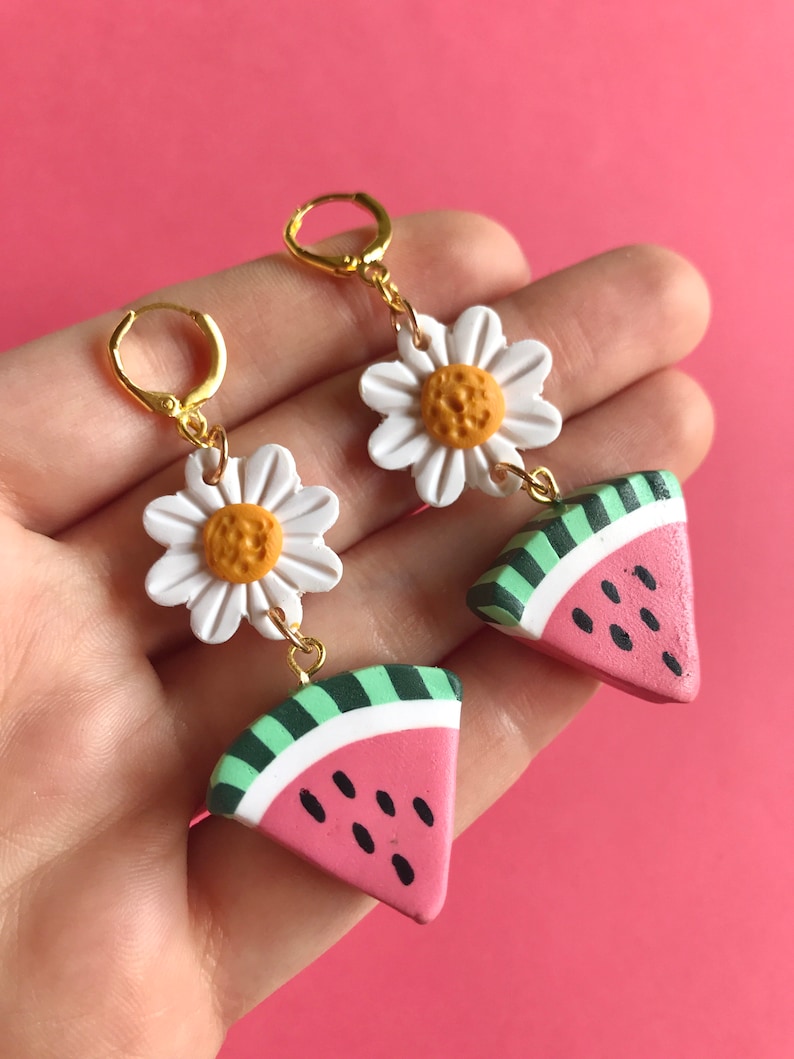 May include: A pair of dangle earrings featuring a white daisy charm and a pink watermelon slice charm. The charms are connected by a gold chain.