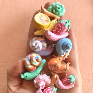 Pu&ograve; includere: Una collezione di ciondoli a forma di lumaca in argilla colorata con vari disegni, tra cui un limone, una fragola, una caramella a bastoncino e pois. I ciondoli sono infilati su catene dorate e sono perfetti per aggiungere un tocco fantasioso a gioielli o artigianato.