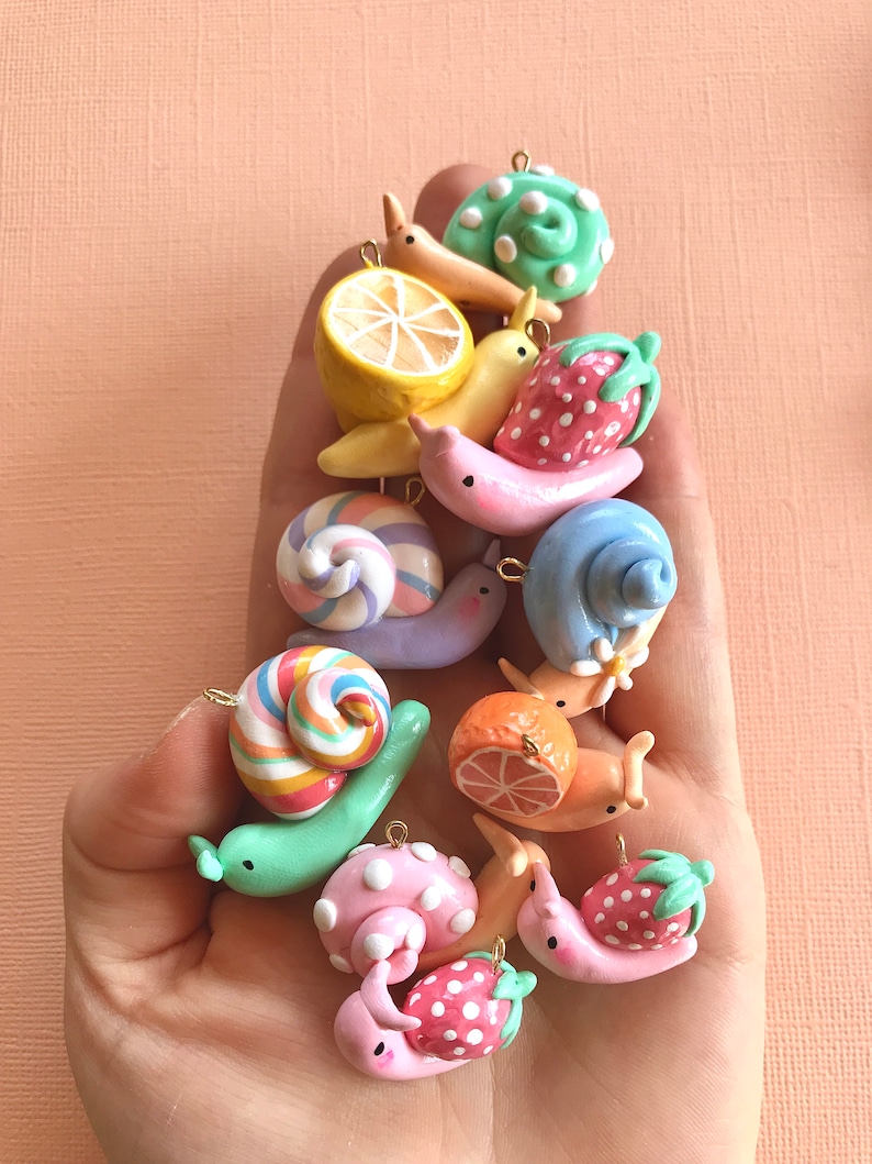 Pu&ograve; includere: Una collezione di ciondoli a forma di lumaca in argilla colorata con vari disegni, tra cui un limone, una fragola, una caramella a bastoncino e pois. I ciondoli sono infilati su catene dorate e sono perfetti per aggiungere un tocco fantasioso a gioielli o artigianato.