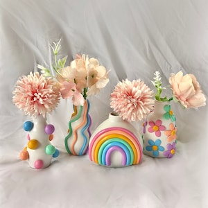 May include: Four pastel-colored ceramic vases with different designs. The vases are filled with pink silk flowers. The vases are on a white background.