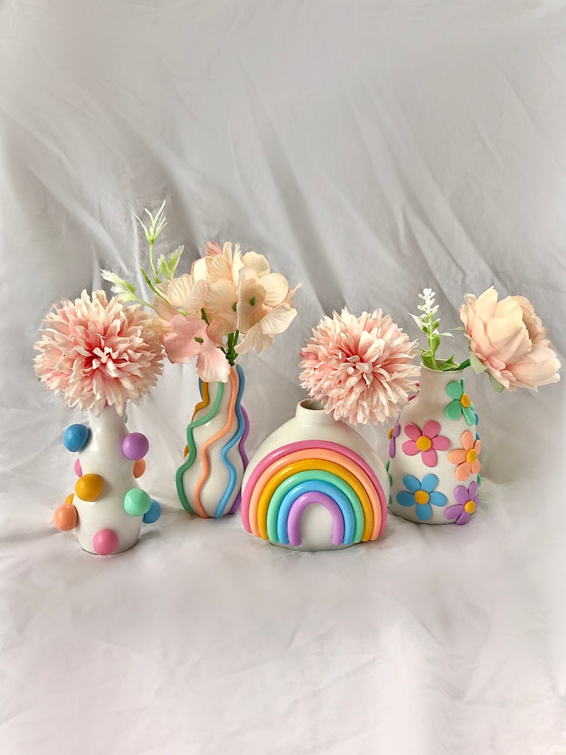 May include: Four pastel-colored ceramic vases with different designs. The vases are filled with pink silk flowers. The vases are on a white background.