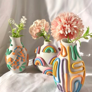 May include: Three ceramic vases with different designs. The vase on the left has a blue and orange pattern. The middle vase has a rainbow pattern. The vase on the right has a multi-colored pattern of lines and curves.