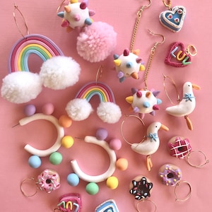 May include: A collection of colorful polymer clay earrings featuring various designs, including rainbows, donuts, hearts, and a spiky ball. The earrings are displayed on a pink background.