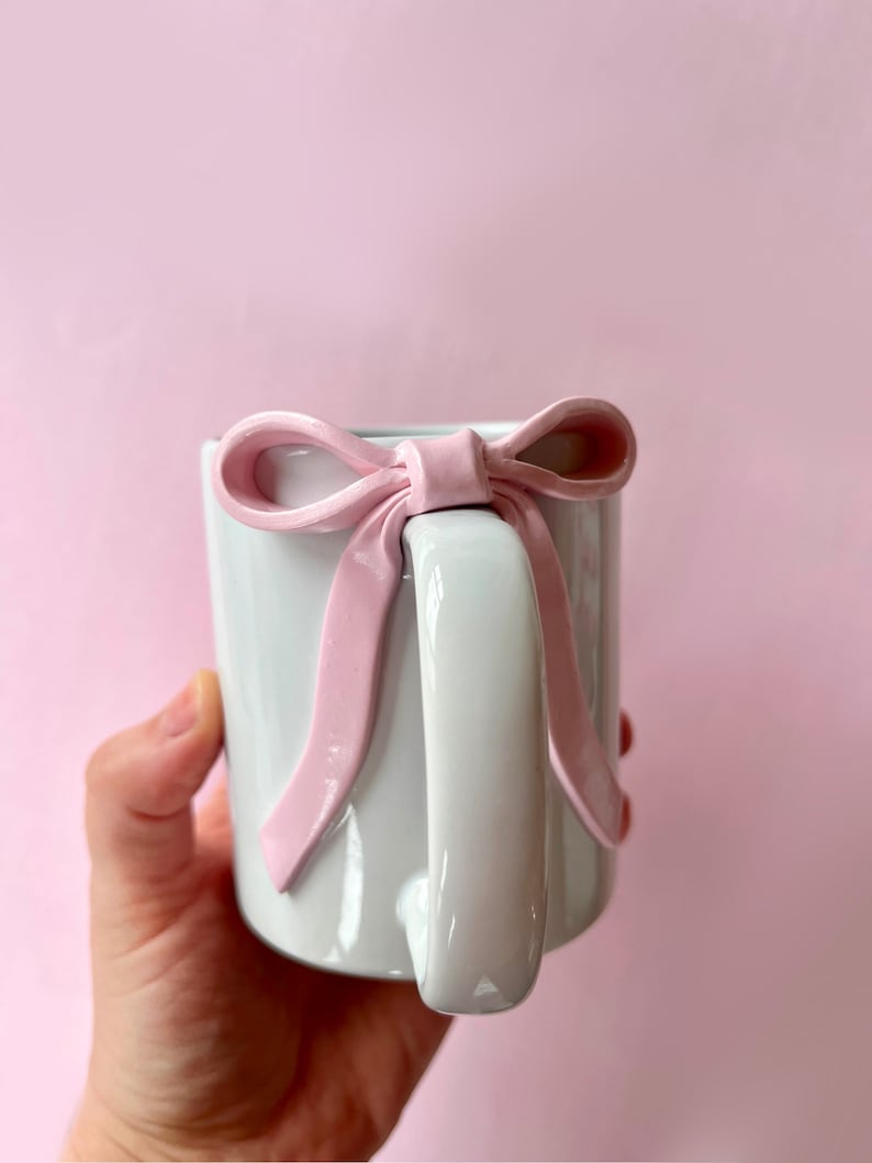 Pu&ograve; includere: Una tazza in ceramica bianca con un grande fiocco rosa attaccato al manico. La tazza &egrave; tenuta da una mano.