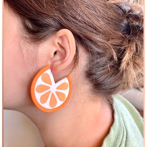 May include: Orange slice shaped earrings with a white center and orange outline. The earrings are worn by a person with brown hair.