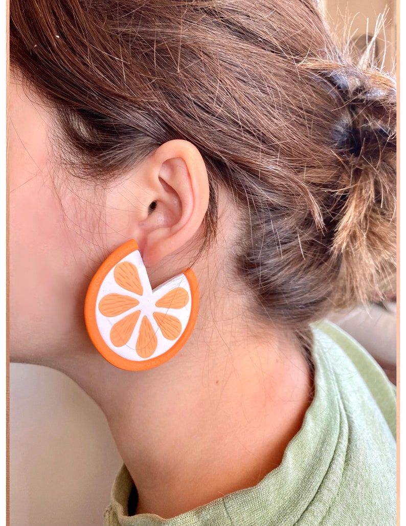 May include: Orange slice shaped earrings with a white center and orange outline. The earrings are worn by a person with brown hair.