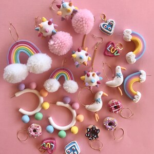 May include: A collection of colorful polymer clay earrings featuring various designs, including rainbows, donuts, hearts, and pom-poms. The earrings are displayed on a pink background.