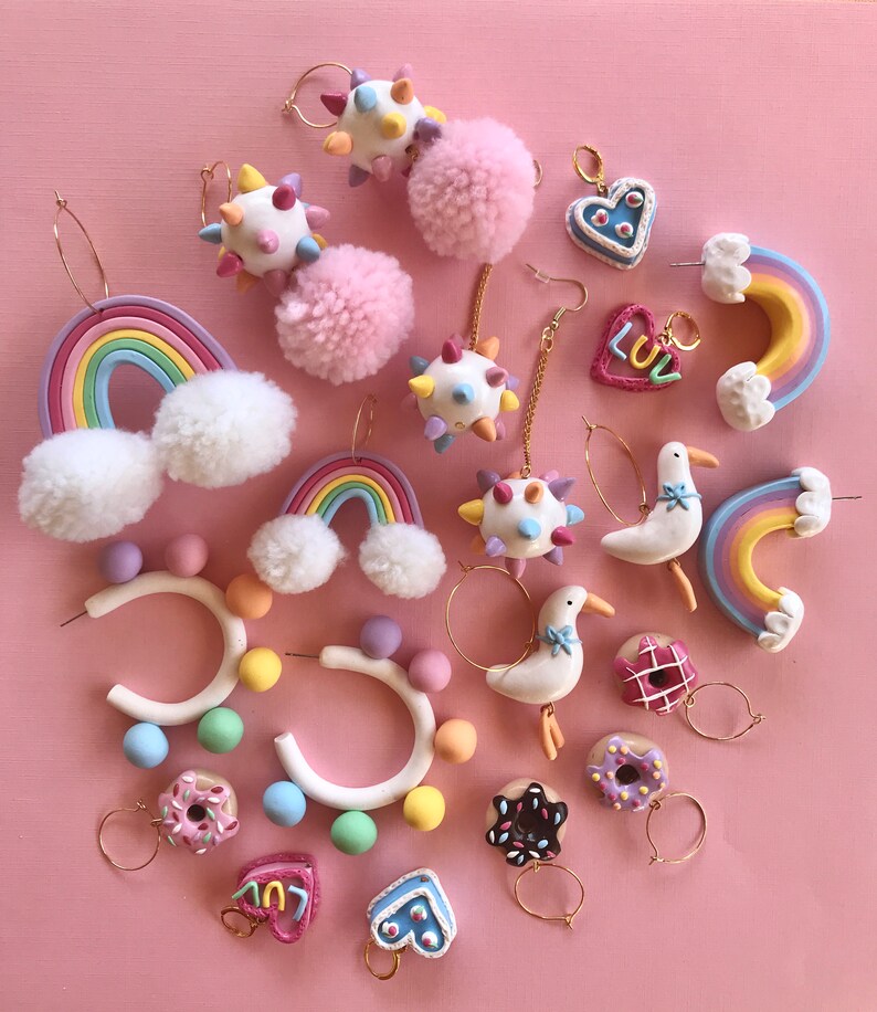 May include: A collection of colorful polymer clay earrings featuring various designs, including rainbows, donuts, hearts, and pom-poms. The earrings are displayed on a pink background.