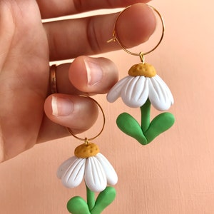 May include: A pair of gold hoop earrings with white daisy flower charms. The flowers have green leaves and yellow centers.