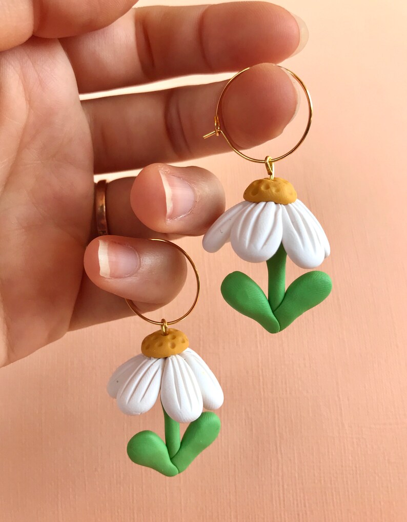 May include: A pair of gold hoop earrings with white daisy flower charms. The flowers have green leaves and yellow centers.