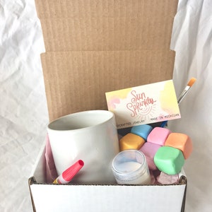 May include: A white cardboard box with a white mug, a red marker, a small jar of white cream, and a set of colorful, soft, square blocks. The box also includes a business card that reads "Sun Sprinkles Handcrafted Jewelry Made in Michigan".