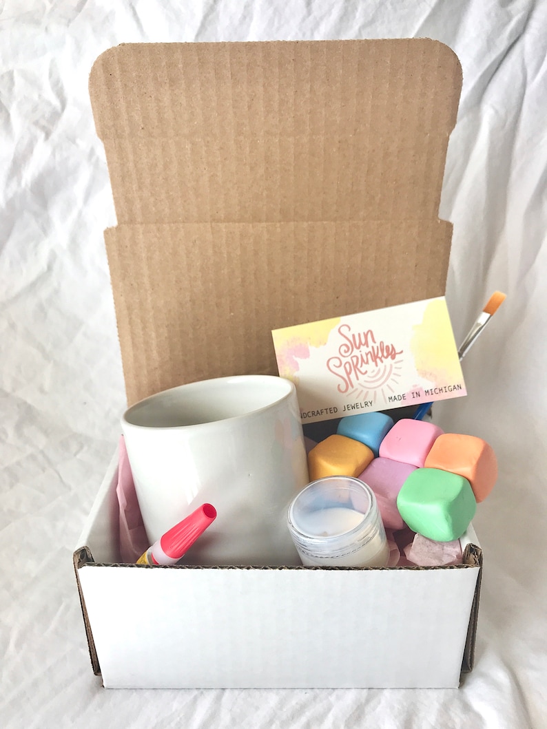 May include: A white cardboard box with a white mug, a red marker, a small jar of white cream, and a set of colorful, soft, square blocks. The box also includes a business card that reads "Sun Sprinkles Handcrafted Jewelry Made in Michigan".