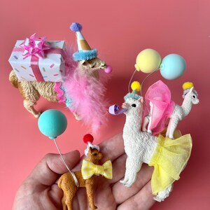 K&ouml;nnte beinhalten: Eine Sammlung skurriler Lama-Figuren, jede mit festlicher Partykleidung. Ein Lama tr&auml;gt ein Geschenk, andere tragen Partyh&uuml;te, Federboas und Luftballons. Die Figuren sind vor einem einfarbigen rosa Hintergrund platziert.