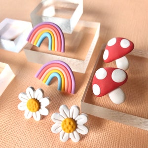 May include: A set of colorful polymer clay earrings featuring a rainbow, a mushroom, and a daisy. The rainbow is made of pink, purple, blue, yellow, and orange clay. The mushroom is red with white polka dots. The daisy is white with a yellow center.