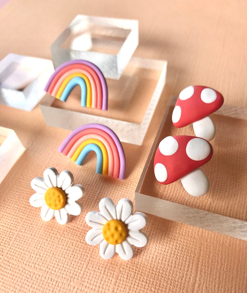 May include: A set of colorful polymer clay earrings featuring a rainbow, a mushroom, and a daisy. The rainbow is made of pink, purple, blue, yellow, and orange clay. The mushroom is red with white polka dots. The daisy is white with a yellow center.
