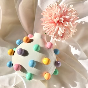 May include: A white ceramic vase with pastel colored polka dots, holding a single pink flower. The vase is round and has a textured surface.