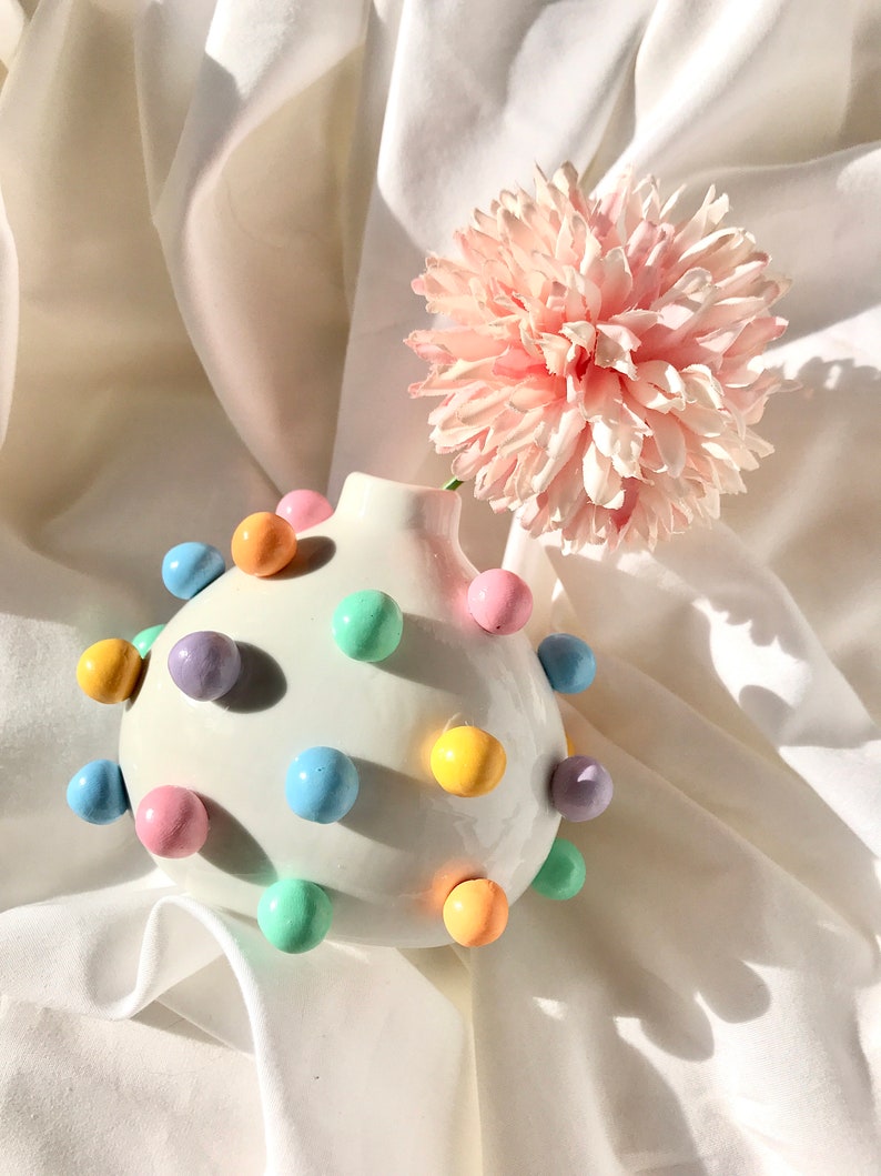 May include: A white ceramic vase with pastel colored polka dots, holding a single pink flower. The vase is round and has a textured surface.