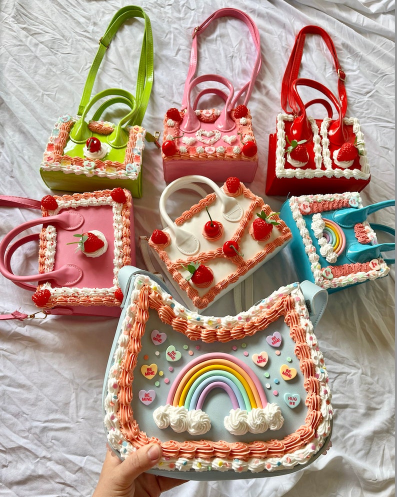 May include: A collection of colorful cake-themed handbags with various designs, including strawberries, cherries, rainbows, and frosting. The handbags have adjustable straps and are made of a glossy material.