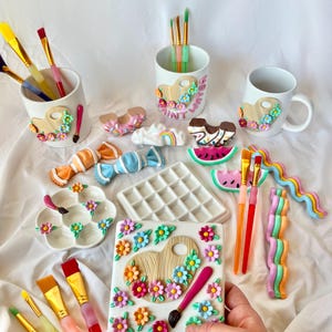 Puede incluir: Un juego de tazas de cerámica blanca con detalles pintados, que incluyen una paleta de pintura, flores y las palabras "Get Your Paint On". Las tazas están llenas de pinceles. También hay varios otros artículos de cerámica pintados, que incluyen una paleta de pintura, flores y una rodaja de sandía.