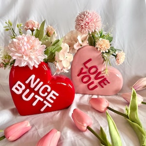 Puede incluir: Dos jarrones de cerámica en forma de corazón, uno rojo con el texto "NICE BUTT" en letras blancas, y otro rosa con el texto "LOVE YOU" en letras rosas. Ambos jarrones están llenos de flores rosas y blancas.