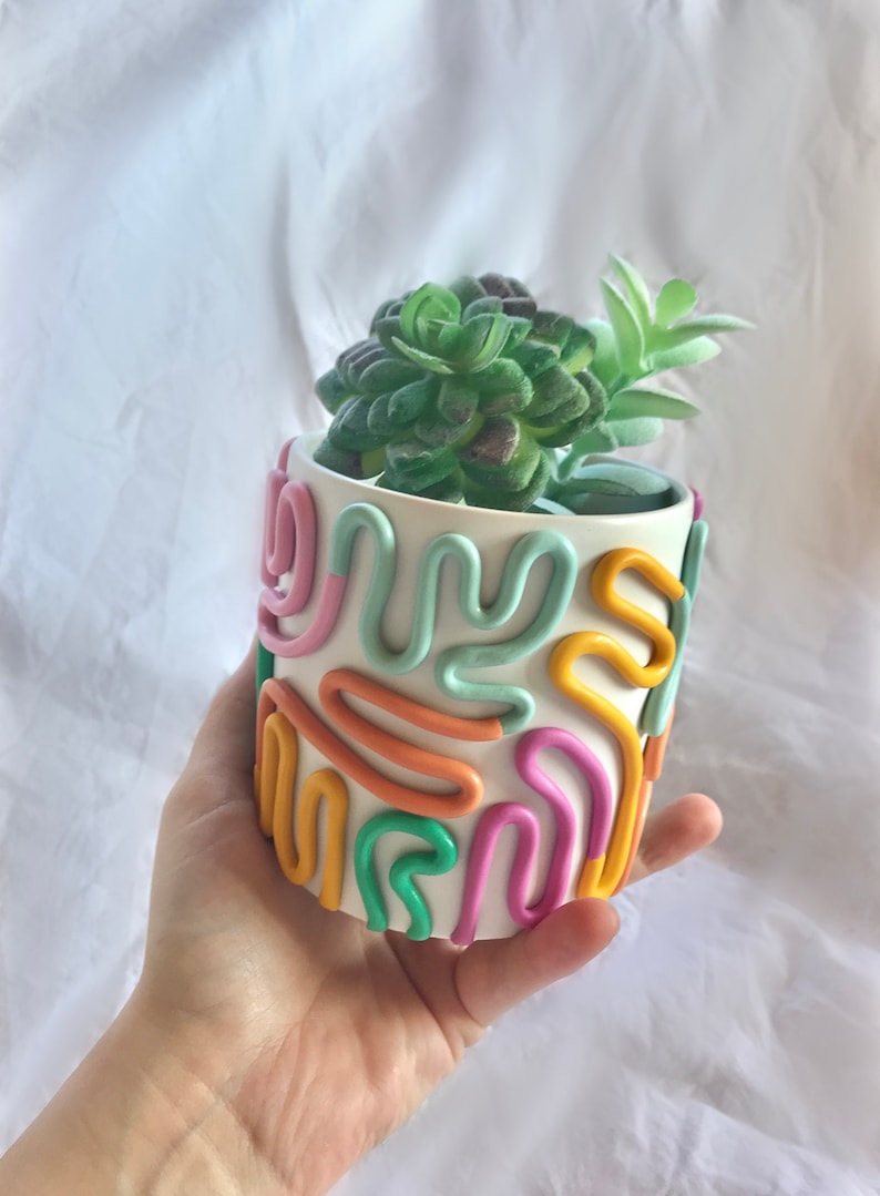 May include: A white ceramic planter with a colourful abstract design. The planter is decorated with pink, orange, yellow, and green lines. A small green succulent plant is growing in the planter.