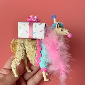K&ouml;nnte beinhalten: Eine skurrile Lama-Figur, geschm&uuml;ckt mit einer Partyh&uuml;tchen, einer rosa Federboa und einer Kaugummiblase. Es tr&auml;gt eine Geschenkbox mit rosa Schleife und bunten Streuseln. Das Lama ist hellbraun und tr&auml;gt ein blau-rosa Outfit.