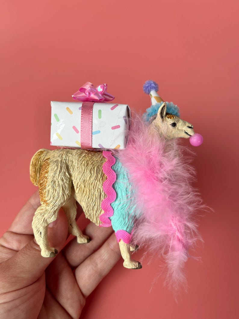 K&ouml;nnte beinhalten: Eine skurrile Lama-Figur, geschm&uuml;ckt mit einer Partyh&uuml;tchen, einer rosa Federboa und einer Kaugummiblase. Es tr&auml;gt eine Geschenkbox mit rosa Schleife und bunten Streuseln. Das Lama ist hellbraun und tr&auml;gt ein blau-rosa Outfit.
