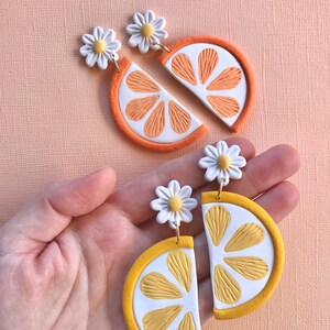 Puede incluir: Pendientes artesanales de arcilla polim&eacute;rica con dise&ntilde;os de rodajas de naranja y lim&oacute;n. Los pendientes de naranja tienen segmentos naranjas y un borde blanco, mientras que los pendientes de lim&oacute;n tienen segmentos amarillos y un borde blanco. Cada pendiente est&aacute; rematado con una margarita blanca.