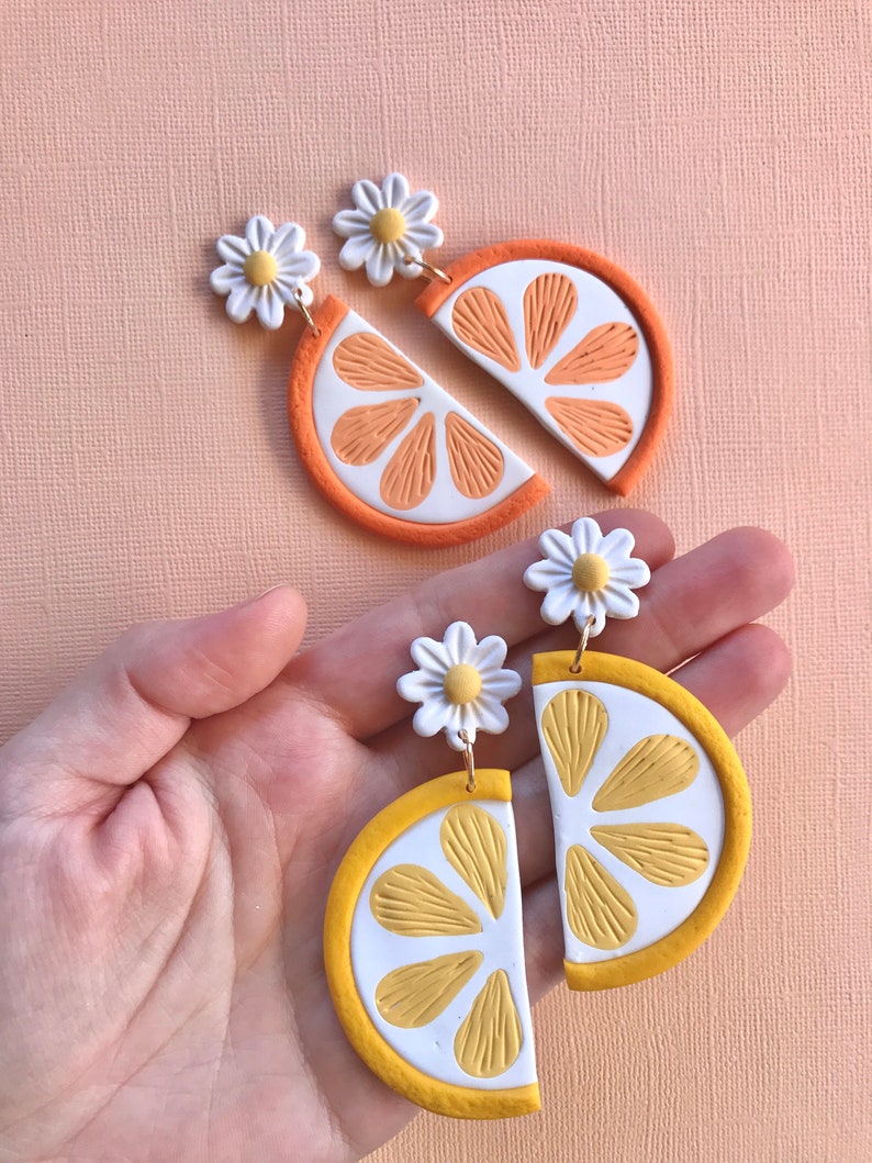 Puede incluir: Pendientes artesanales de arcilla polim&eacute;rica con dise&ntilde;os de rodajas de naranja y lim&oacute;n. Los pendientes de naranja tienen segmentos naranjas y un borde blanco, mientras que los pendientes de lim&oacute;n tienen segmentos amarillos y un borde blanco. Cada pendiente est&aacute; rematado con una margarita blanca.