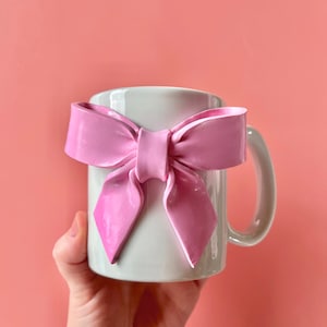 Pu&ograve; includere: Una tazza in ceramica bianca con un fiocco rosa attaccato al manico. La tazza &egrave; tenuta da una mano.