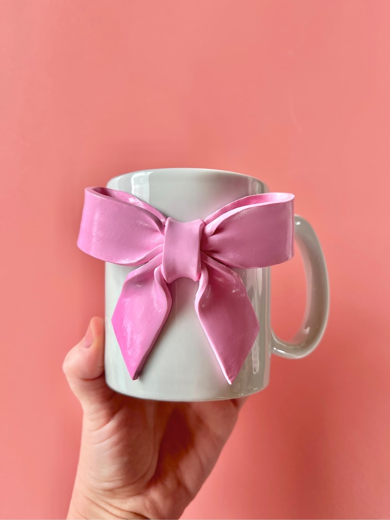 Pu&ograve; includere: Una tazza in ceramica bianca con un fiocco rosa attaccato al manico. La tazza &egrave; tenuta da una mano.