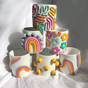 May include: A stack of six white ceramic planters with colourful, hand-painted designs. The designs include rainbows, flowers, smiley faces, and abstract patterns.