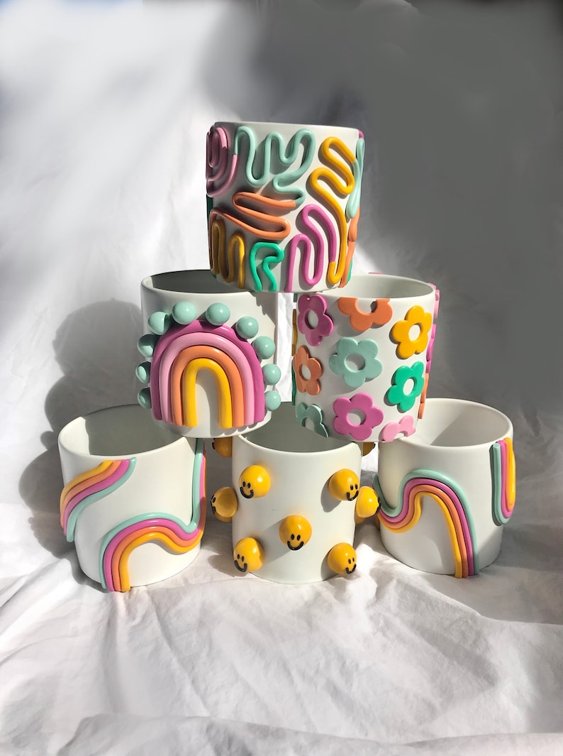 May include: A stack of six white ceramic planters with colourful, hand-painted designs. The designs include rainbows, flowers, smiley faces, and abstract patterns.