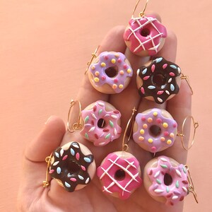 May include: Eight donut-shaped earrings with different colored glazes and sprinkles. The earrings are made of polymer clay and have gold hoops.