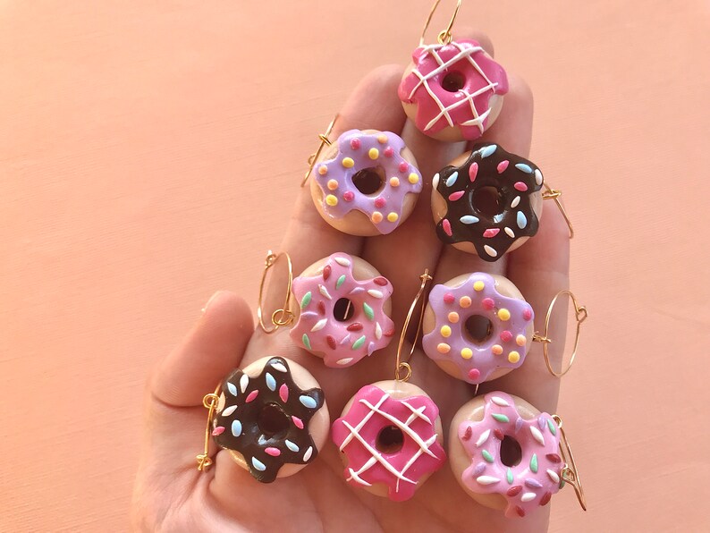 May include: Eight donut-shaped earrings with different colored glazes and sprinkles. The earrings are made of polymer clay and have gold hoops.
